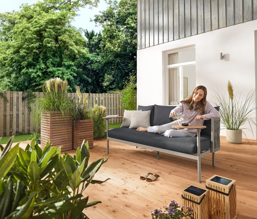 Eine Frau gießt draußen auf einer Gartenterrasse Wasser in ein Glas, neben ihr stehen eine große und eine kleine Bambus-Solar-Laterne, sowie 5 LED-Solar-Spieße und eine Gartensofa mit 2 Drehtischen. Außerdem sind 2 Pflanzsäulen »Tinus« zu sehen.