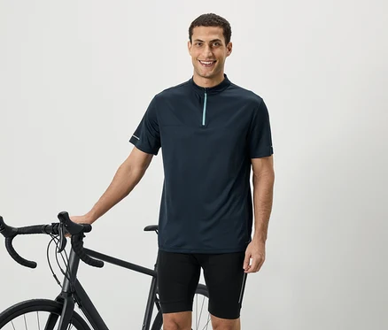 Ein lächelnder Mann in einem marineblauen Kurzarmhemd mit Reißverschluss steht neben einem Fahrrad.