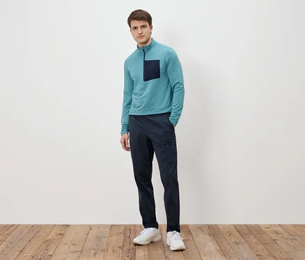 Mann steht vor einer weißen Wand, trägt ein türkisfarbenes Langarmshirt mit dunkler Tasche, marineblaue Hose und weiße Turnschuhe.