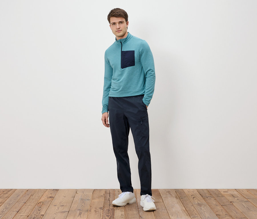 Mann steht vor einer weißen Wand, trägt ein türkisfarbenes Langarmshirt mit dunkler Tasche, marineblaue Hose und weiße Turnschuhe.