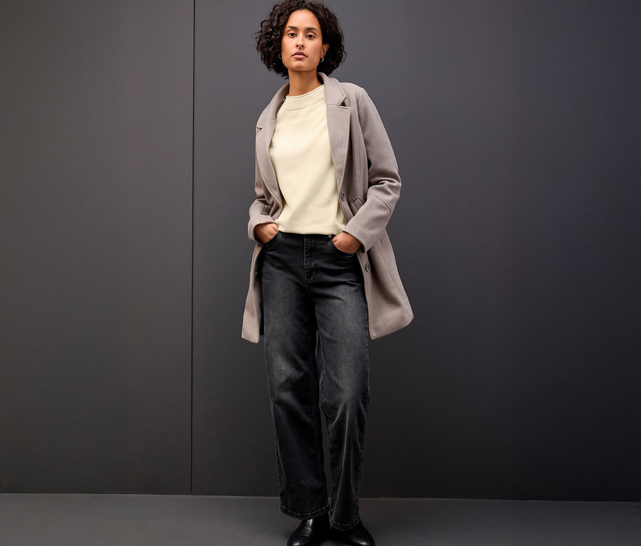 Frau steht mit den Händen in den Hosentaschen und trägt einen beigen Street One Strickpullover mit Rollkante, eine graue Jacke und eine dunkle Jeans.