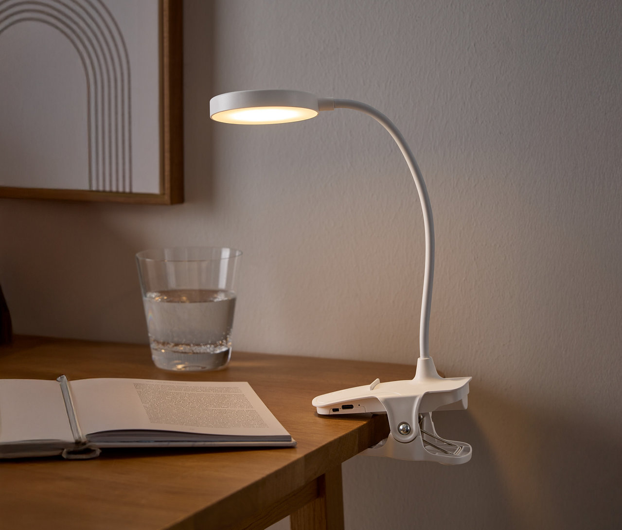 Eine weiße Klemmlampe beleuchtet ein offenes Buch auf einem Holztisch, neben einem Glas Wasser und einem gerahmten Bild.