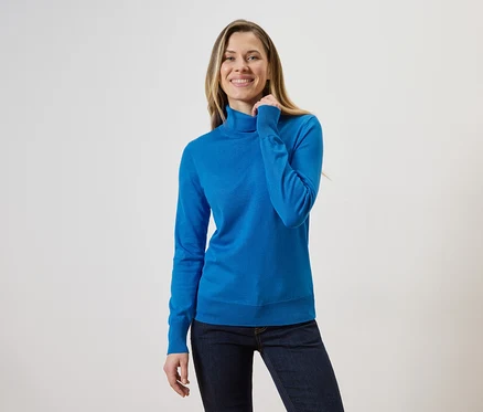 Frau mit blonden Haaren, die einen blauen Rollkragenpullover und Jeans vor einem hellen Hintergrund trägt.