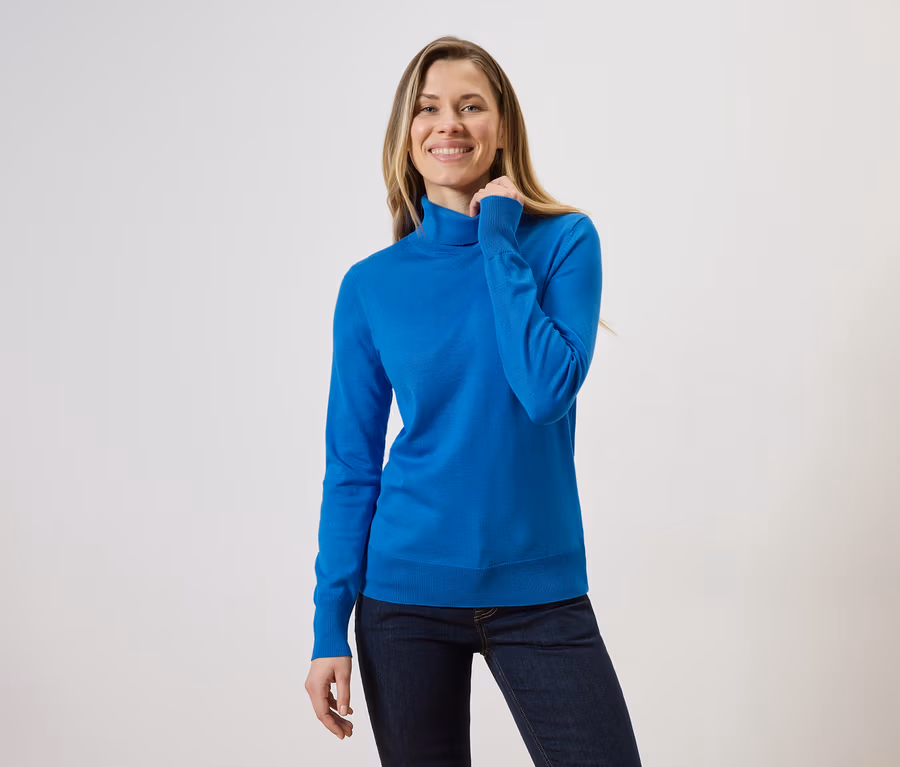 Frau mit blonden Haaren, die einen blauen Rollkragenpullover und Jeans vor einem hellen Hintergrund trägt.