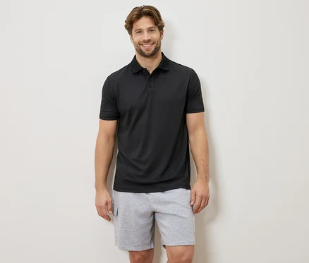 Ein lächelnder Mann steht vor einer Wand und trägt ein schwarzes Poloshirt und graue Shorts.