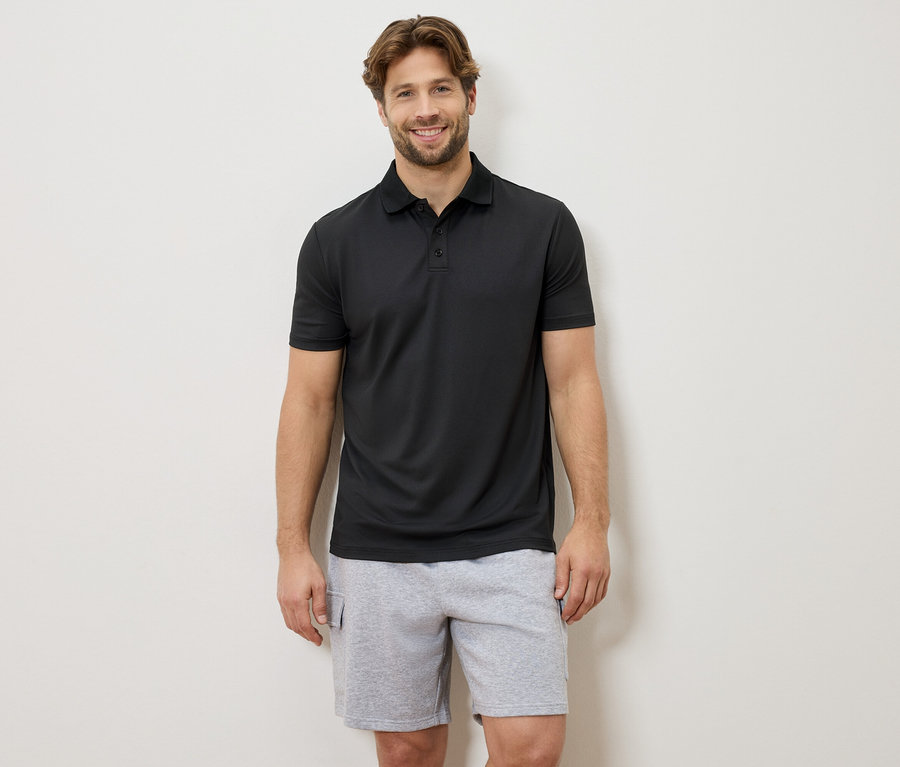 Ein lächelnder Mann steht vor einer Wand und trägt ein schwarzes Poloshirt und graue Shorts.