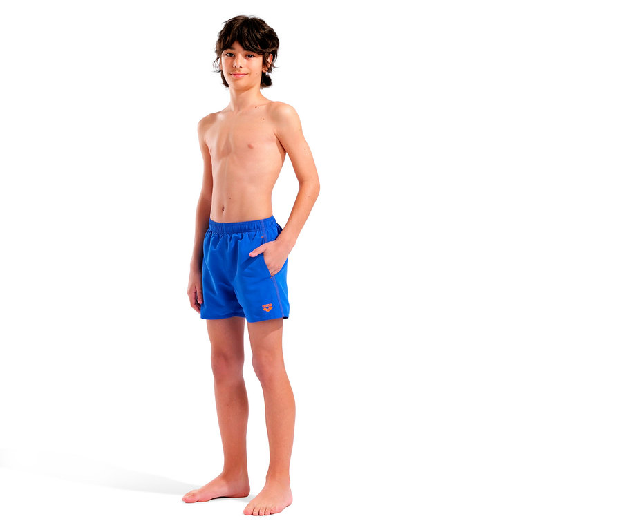 Ein junger, barfüßiger Junge mit braunen Haaren steht in blauen Badeshorts mit orangenem Logo auf weißem Hintergrund.