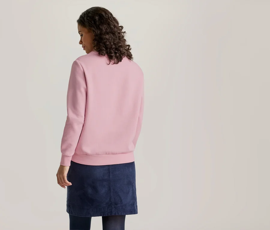 Eine Frau mit dunklem, lockigem Haar steht mit dem Rücken zum Betrachter und blickt leicht nach rechts. Sie trägt ein schlichtes rosa Langarm-Sweatshirt und einen dunkelblauen Rock.