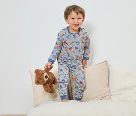 Ein lächelnder Junge im blauen Schlafanzug mit Baufahrzeugmuster steht mit einem Teddybär auf einem Sofa.