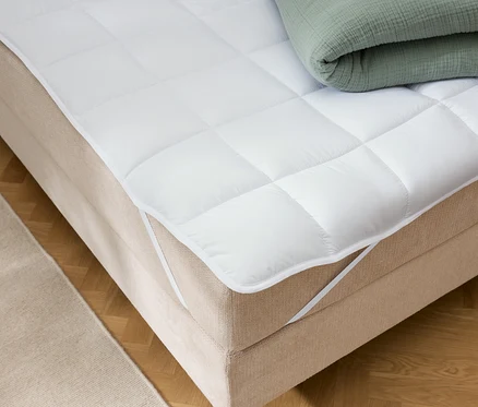 Eine weiße, wattierte Matratzenauflage mit Gummibändern an den Ecken liegt auf einem beigen Bett. Eine gefaltete grüne Decke liegt obenauf.