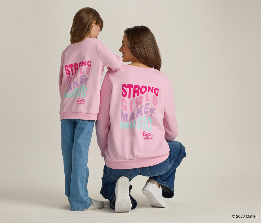 Rückansicht einer jungen Frau, die neben einem jungen Mädchen kniet. Beide tragen hellrosa Sweatshirts mit dem Aufdruck "Strong Girls Make Music".