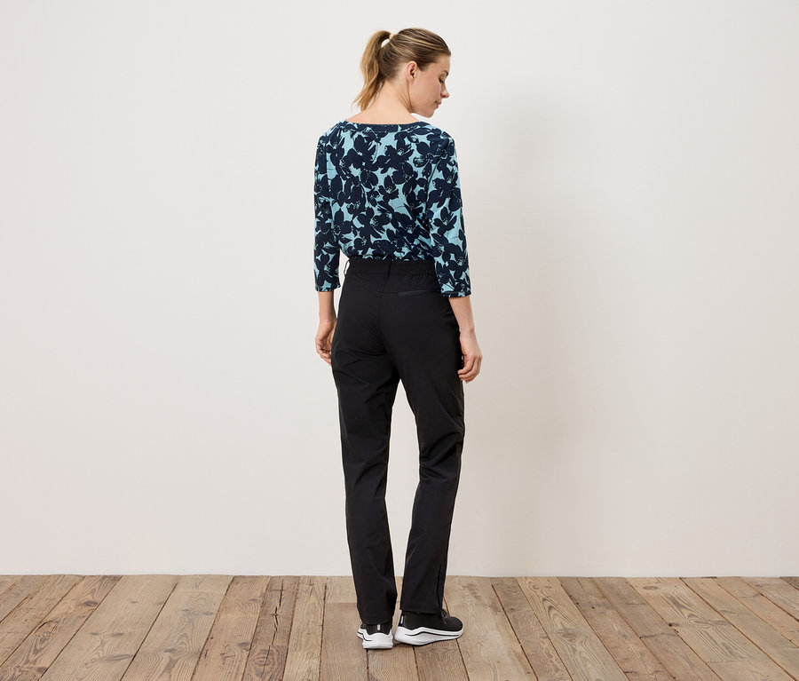 Eine Frau steht mit dem Rücken zur Kamera und trägt eine marineblaue Bluse mit Blumenmuster und eine schwarze Hose.