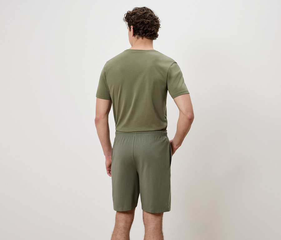 Rückansicht eines Mannes mit olivgrünem T-Shirt und Shorts, die Hände in den Hosentaschen.