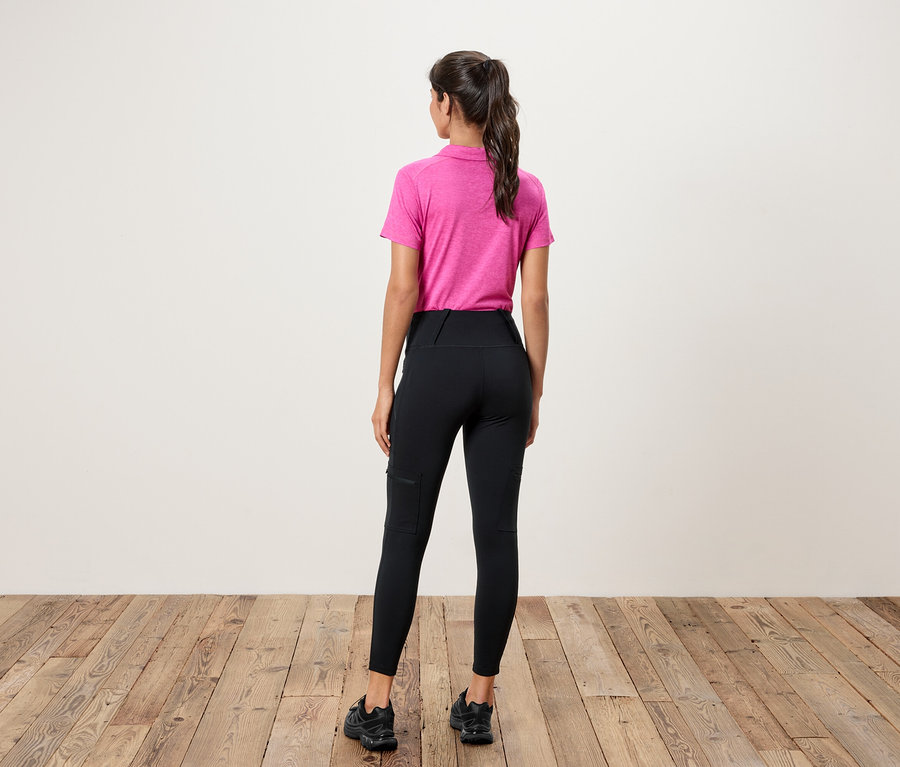 Eine Frau steht mit dem Rücken zur Kamera und trägt ein pinkes Polohemd und schwarze Leggings mit Taschen.