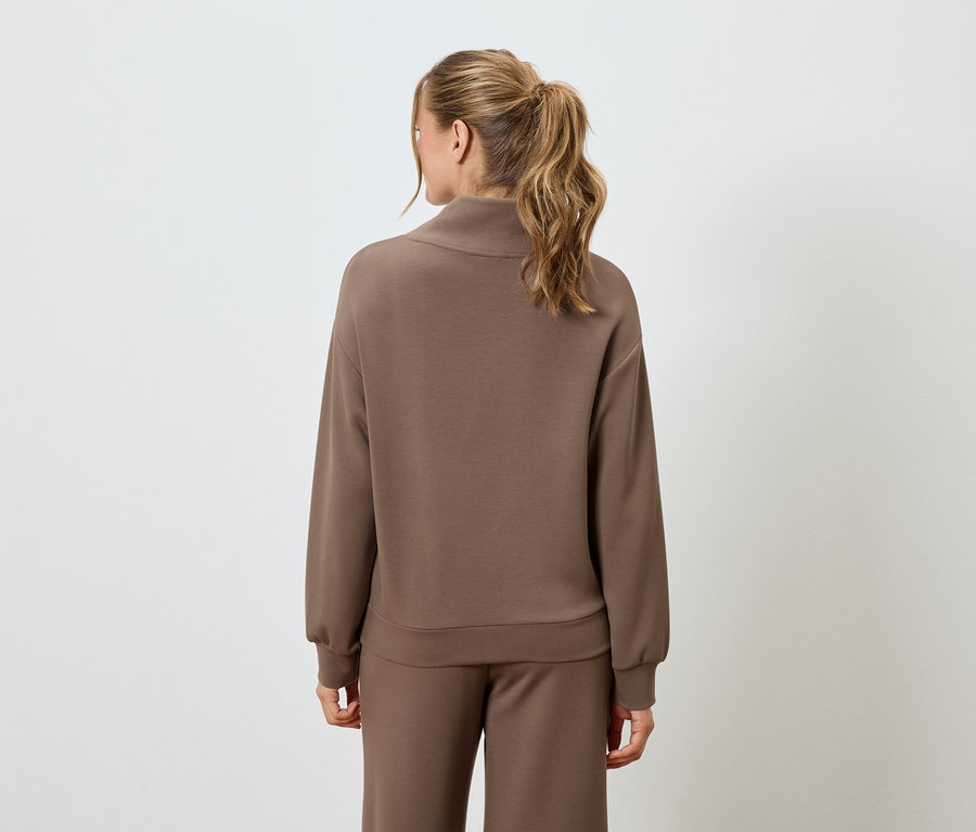 Rückansicht einer Frau mit blonden Haaren, die einen braunen Pullover und eine braune Hose trägt.