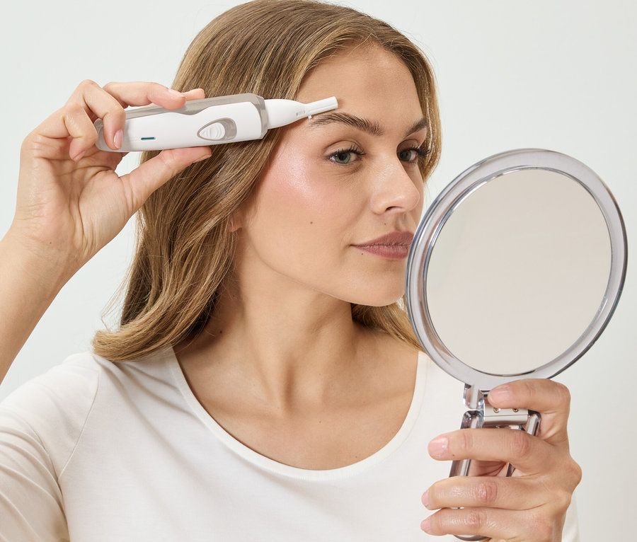 Frau zupft ihre Augenbrauen mit einem weißen Epilierer, während sie in einen runden Spiegel schaut.