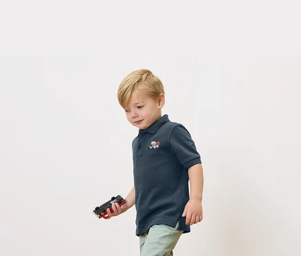 Ein blonder Junge hält ein Spielzeugauto in der Hand und trägt ein marineblaues Poloshirt mit einer kleinen Zugapplikation.