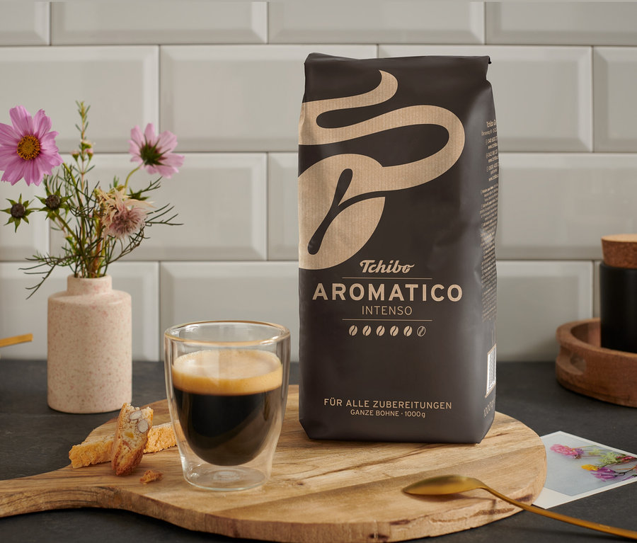 Eine Tasse Kaffee steht neben einer Packung Aromatico Intenso - 1 kg Ganze Bohne auf einem Holztisch.