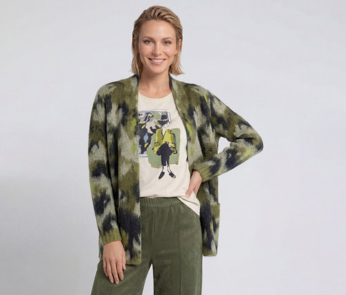 Lächelnde Frau mit kurzen blonden Haaren trägt eine grün gemusterte Strickjacke, ein helles T-Shirt mit Grafikdruck und olivgrüne Hosen.