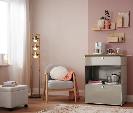 Wohnzimmer mit Sessel, Lampe und Kommode mit Kaffeemaschine. Auf der Kommode steht ein Kapselkorb, weiß.