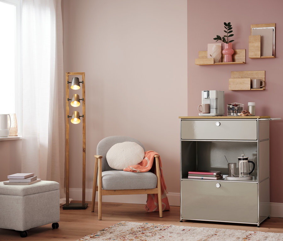 Wohnzimmer mit Sessel, Lampe und Kommode mit Kaffeemaschine. Auf der Kommode steht ein Kapselkorb, weiß.