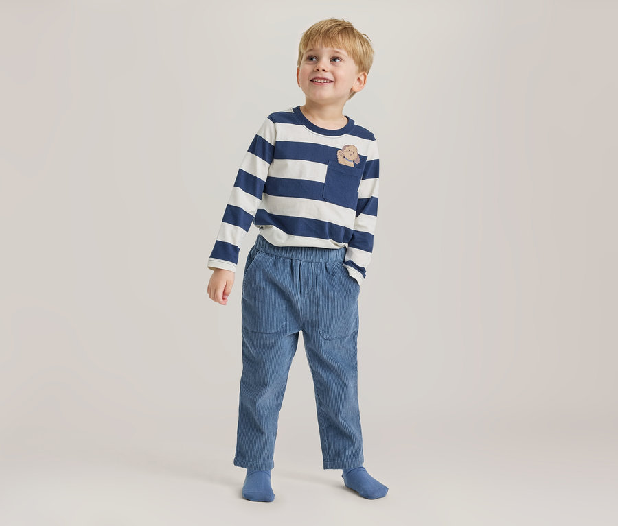 Junger Junge steht, blickt lächelnd nach oben und trägt ein blau-weiß gestreiftes Langarmshirt mit Tier-Brusttasche, blaue Cordhose und blaue Socken.