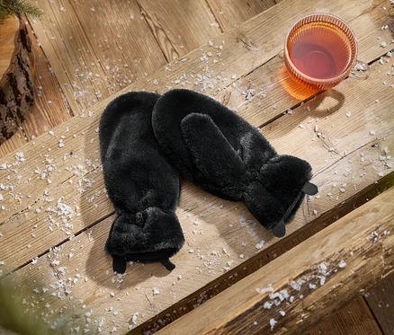 Ein Paar schwarze Kunstfell-Fäustlinge neben einer Tasse Tee auf einer schneebedeckten Holzoberfläche mit einem Tannenzapfen und Tannenzweig.