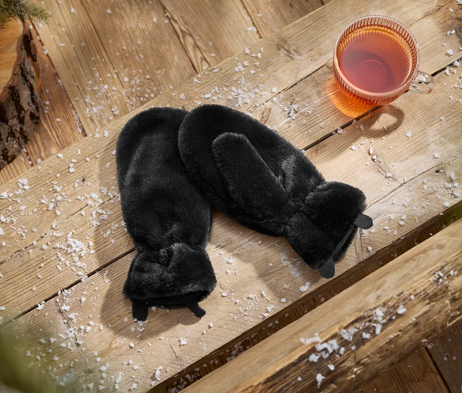 Ein Paar schwarze Kunstfell-Fäustlinge neben einer Tasse Tee auf einer schneebedeckten Holzoberfläche mit einem Tannenzapfen und Tannenzweig.