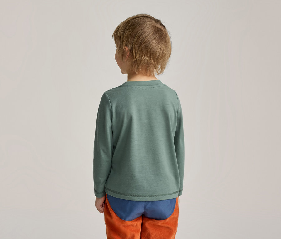 Ein Kind von hinten gesehen, das ein grünes Langarmshirt, blaue Unterwäsche und eine orangefarbene Hose trägt.