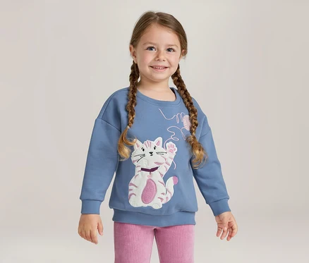 Lächelndes junges Mädchen mit geflochtenen Zöpfen, das ein blaues Sweatshirt mit einer weiß-rosa Katzenapplikation und eine rosa Ripphose trägt.