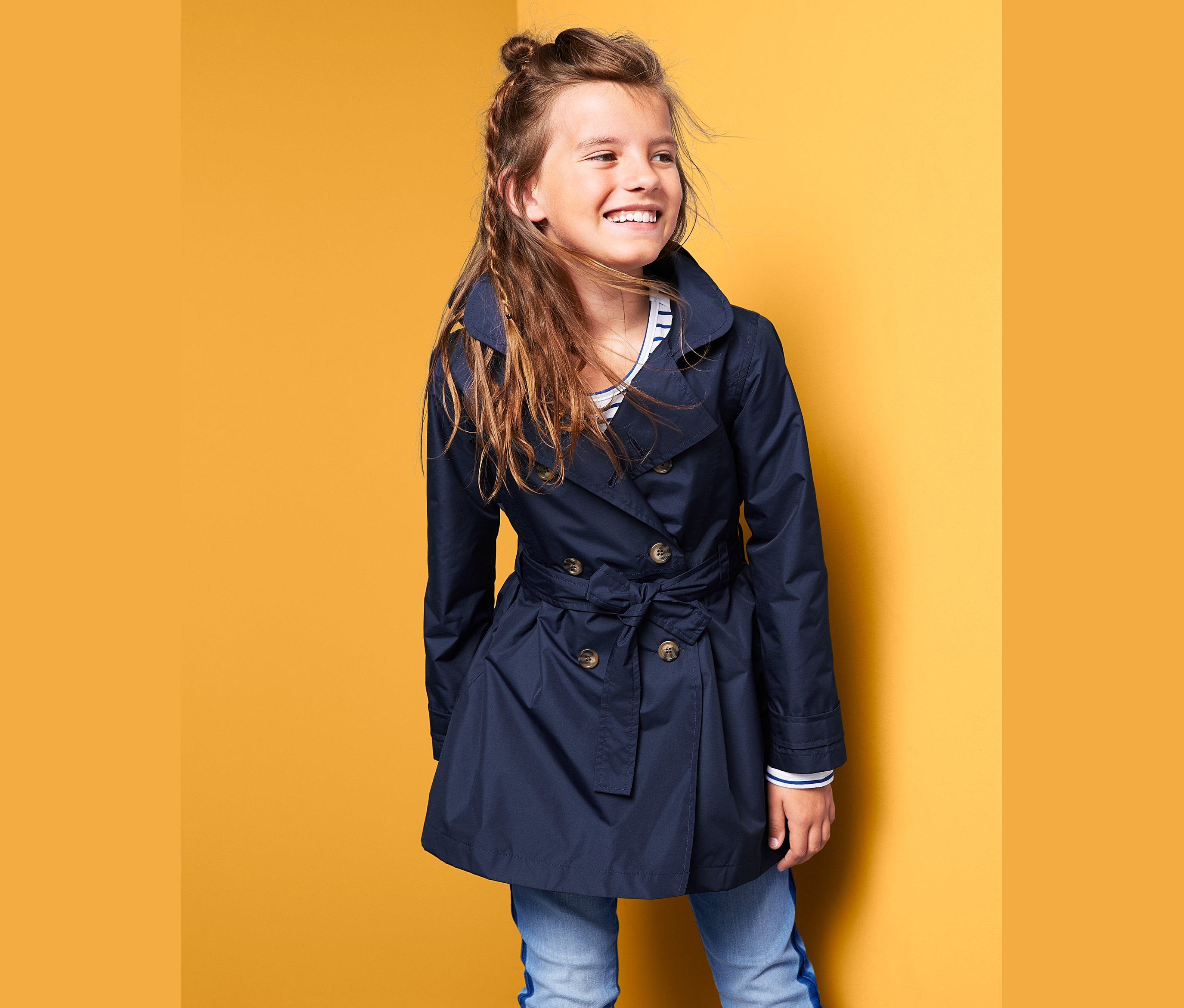 Lächelndes Mädchen posiert in einem dunkelblauen Allwetter-Trenchcoat.