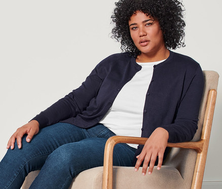 Eine Frau sitzt in einem Sessel und trägt einen dunkelblauen Feinstrick-Cardigan, ein weißes Shirt mit 3/4-Arm, eine dunkelblaue Slim-Denim »Fit Emma« und Leder-Loafer.