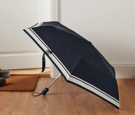 Geöffneter Automatik-Taschenschirm liegt auf Holzboden neben Fußmatte und einem Paar Schuhe.