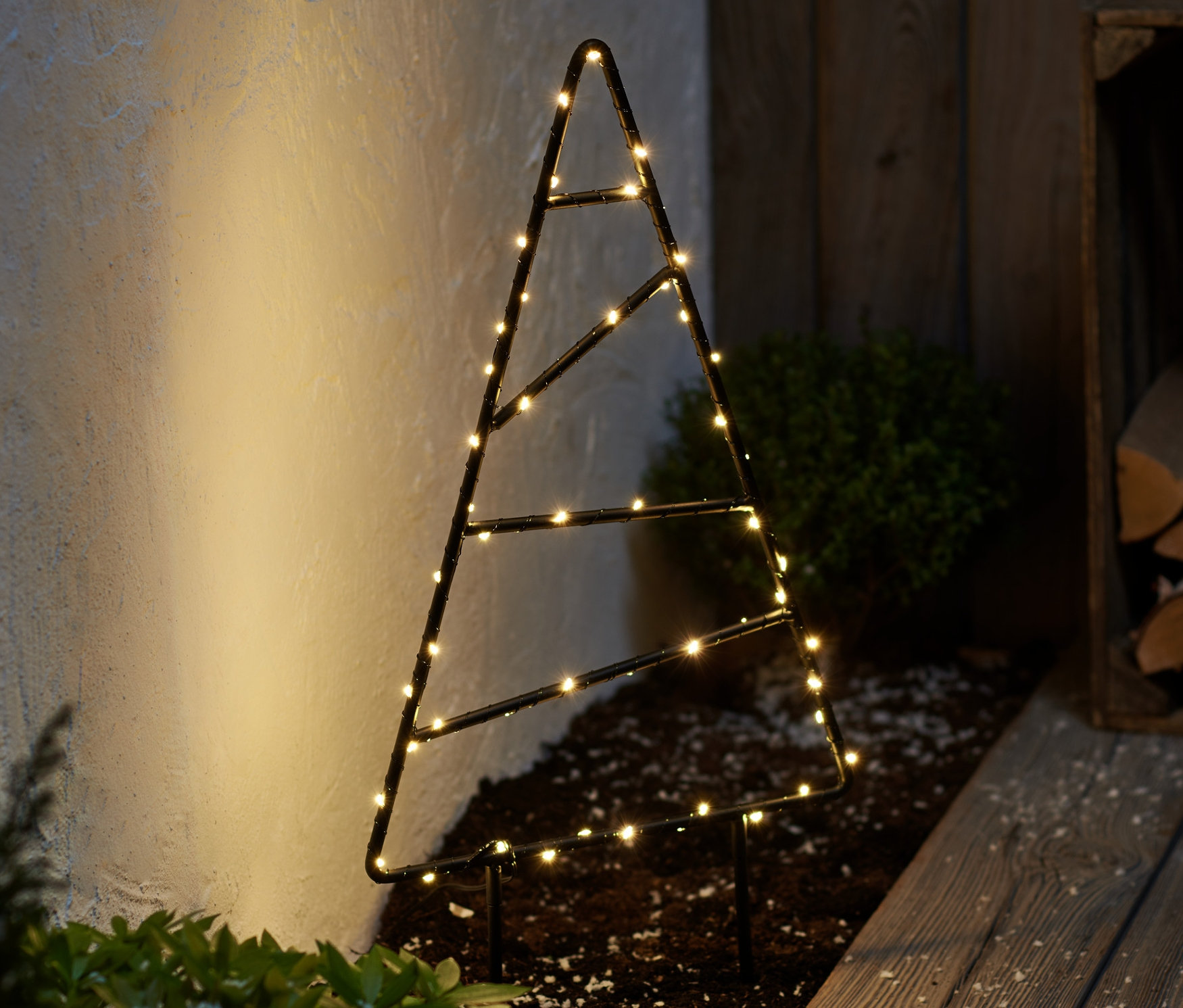 Eine kleine LED-Outdoor-Tanne aus Metall steht auf dem Boden neben einem Holzsteg.