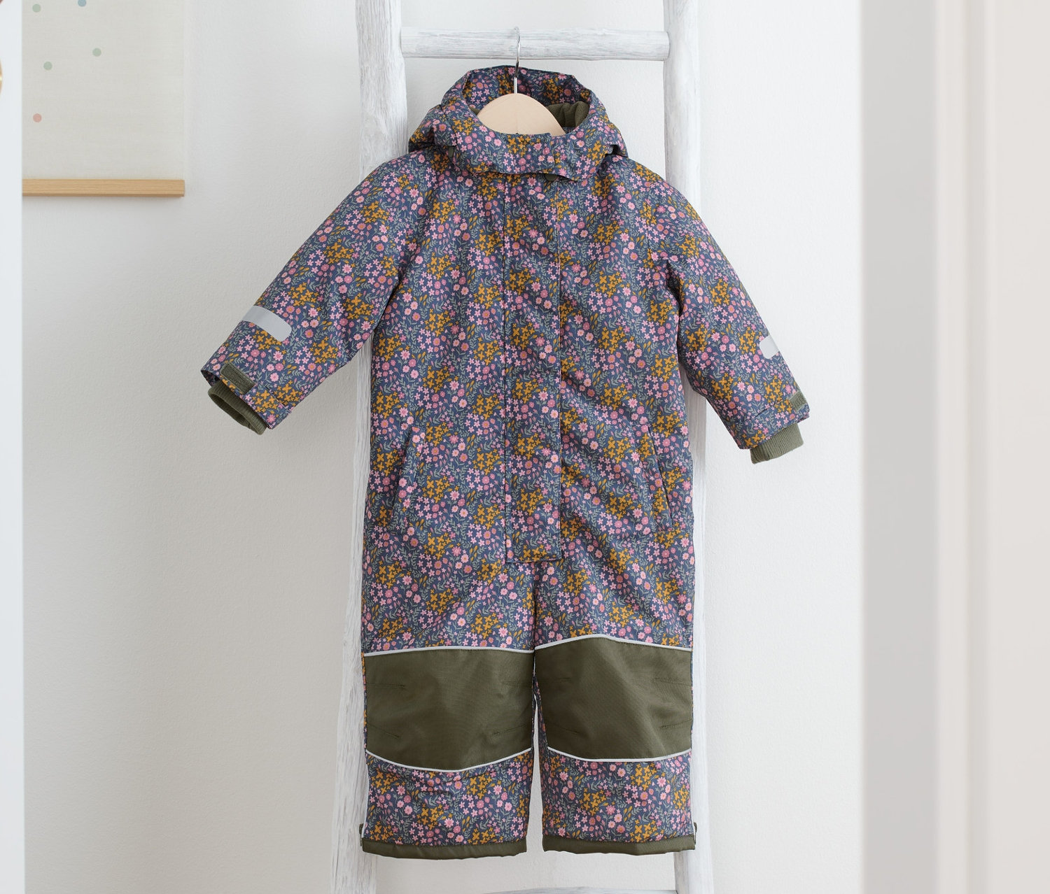 Ein Schneeanzug mit Blümchen-Print hängt auf einer weißen Leiter, daneben liegen gelbe Stiefel.