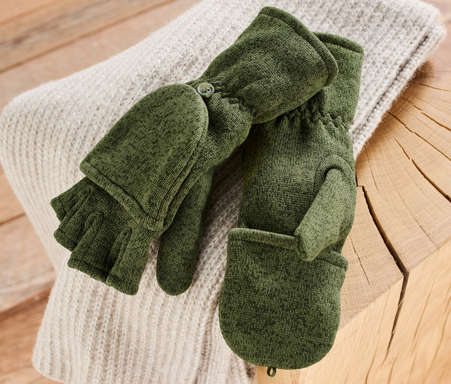 Khakifarbene Strickfleece-Klappfäustlinge liegen auf einem Pullover auf einem Holztisch.