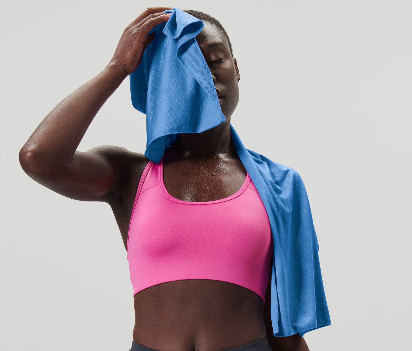 Frau wischt sich mit einem himmelblauen, kühlenden Sporthandtuch das Gesicht. Sie trägt einen rosa Sport-BH und dunkelgraue Leggings.