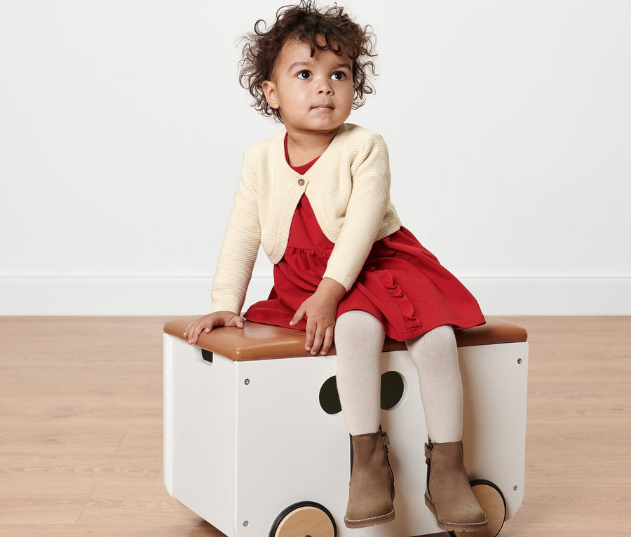 Mädchen sitzt auf Spielzeugkiste, trägt Baby-Strickbolero.