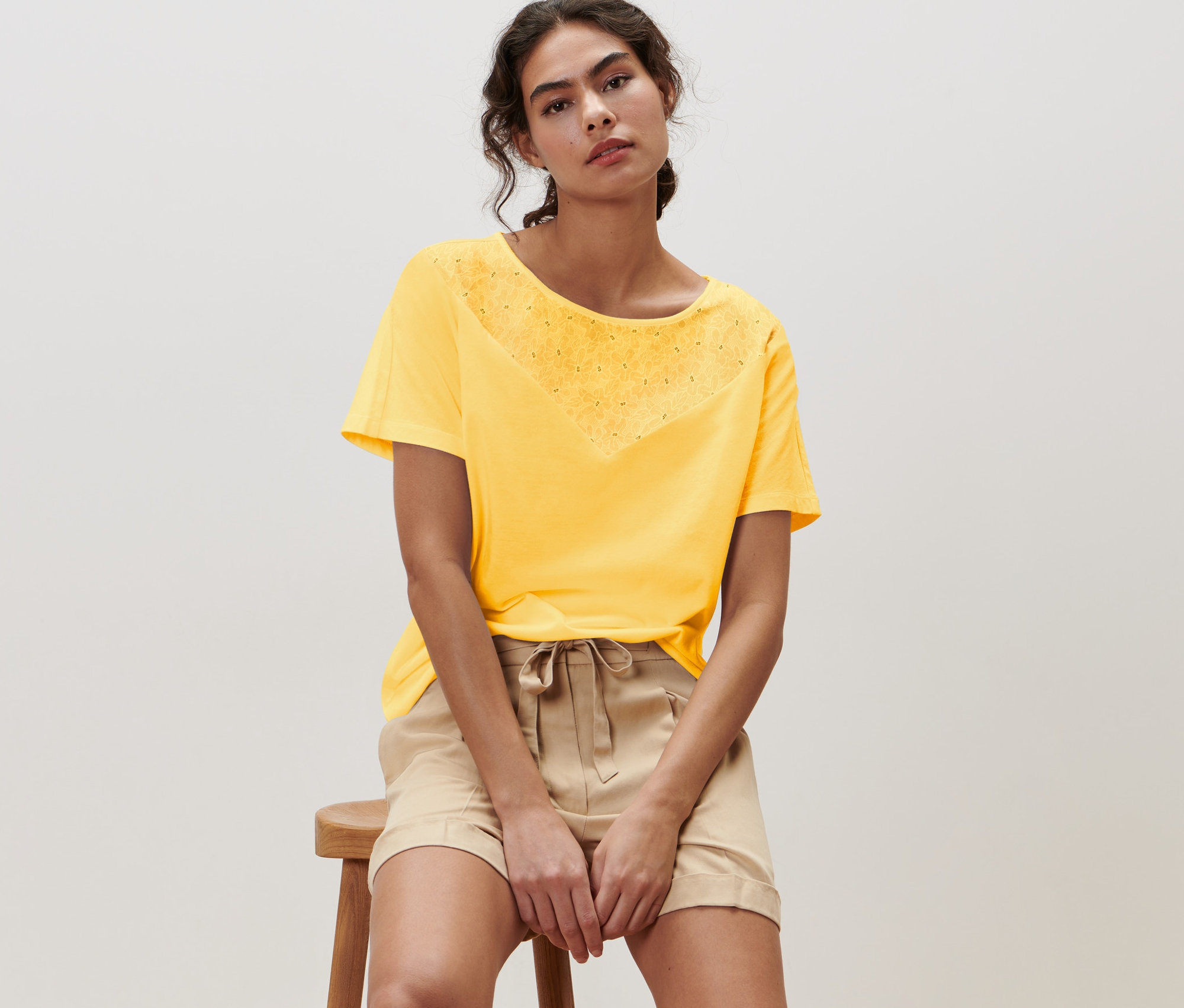Frau sitzt auf einem Hocker und trägt ein gelbes Blusenshirt mit Stickerei.