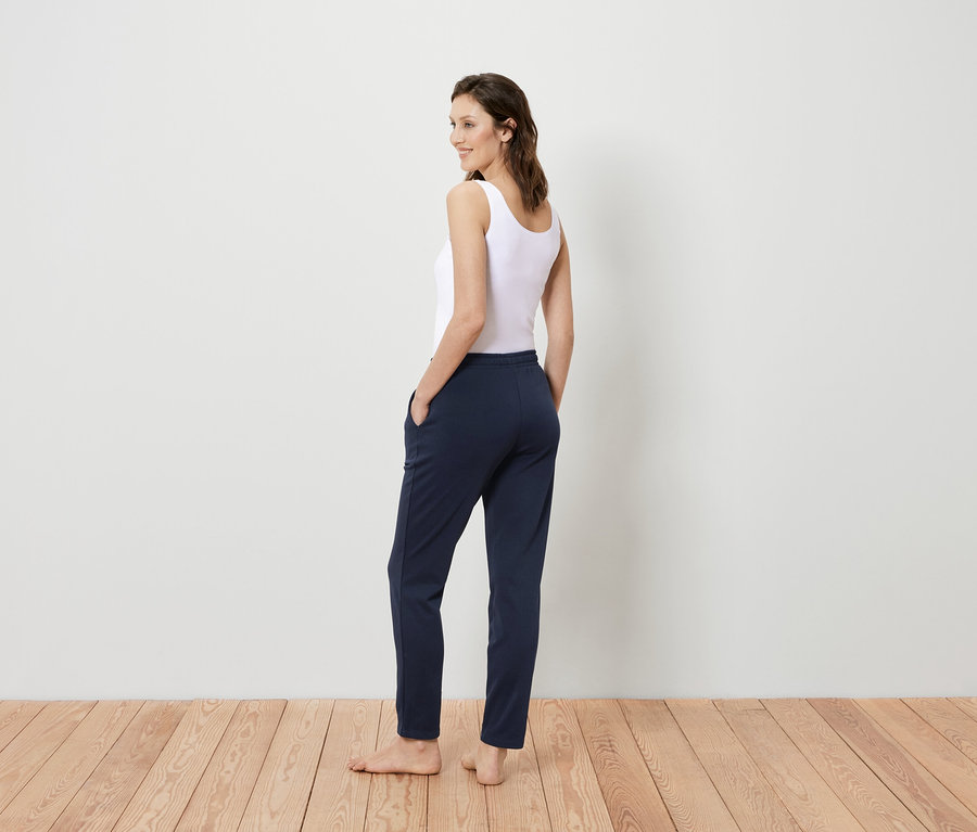 Frau in weißem Tanktop und dunkelblauer Hose steht mit dem Rücken zur Kamera und blickt über die Schulter.