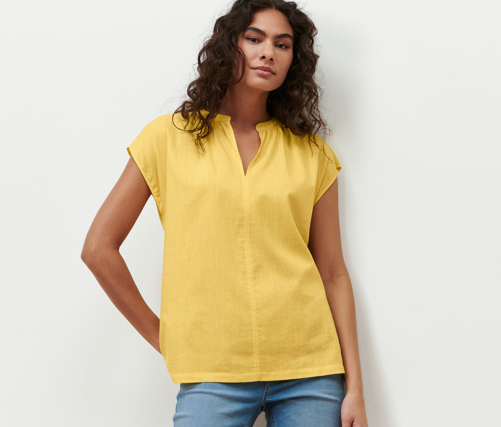 Frau posiert in gelber Kurzarmbluse und blauen Jeans.