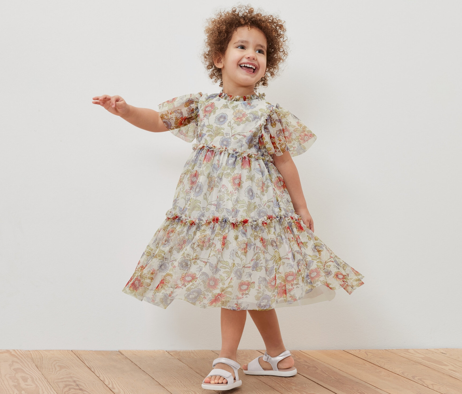 Ein lächelndes Mädchen posiert in einem geblümten Kinder-Tüllkleid und weißen Sandalen.