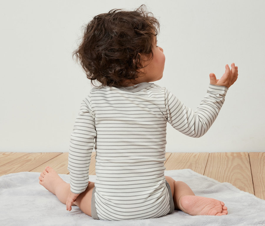 Ein Kind sitzt mit dem Rücken zur Kamera auf einer Unterlage und trägt einen 5er-Pack Baby-Bodys mit Mitwachsfunktion.