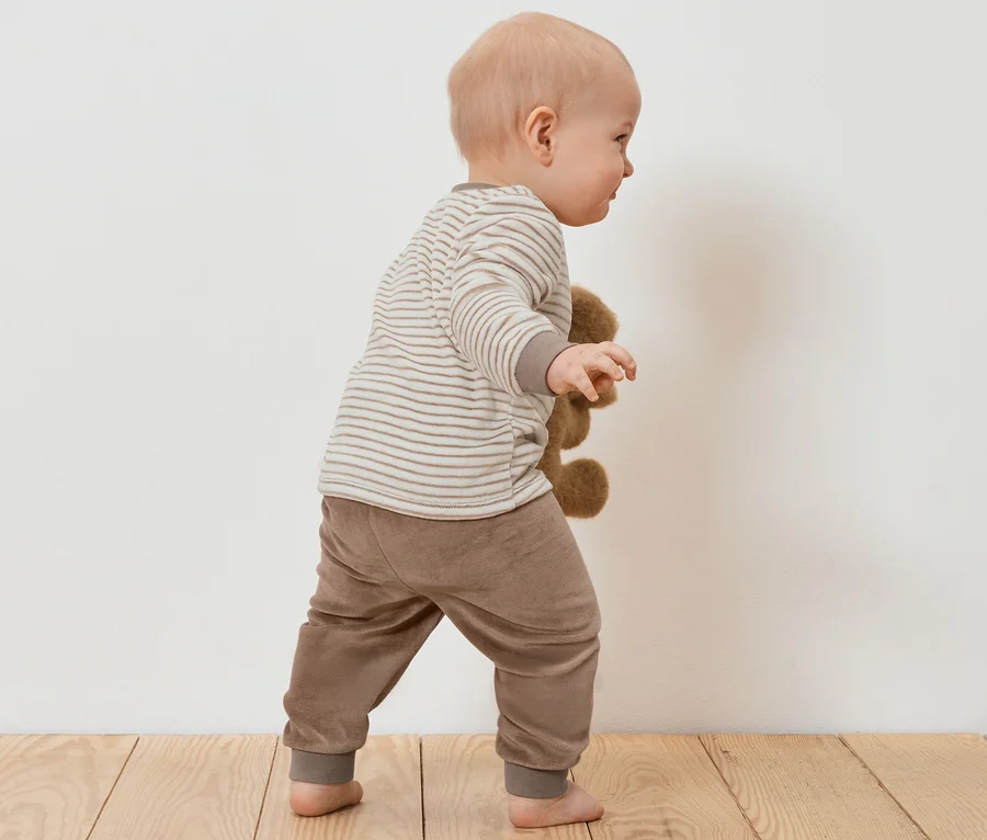 Kleinkind im Baby-Nicki-Set hält Teddybär auf Holzboden.