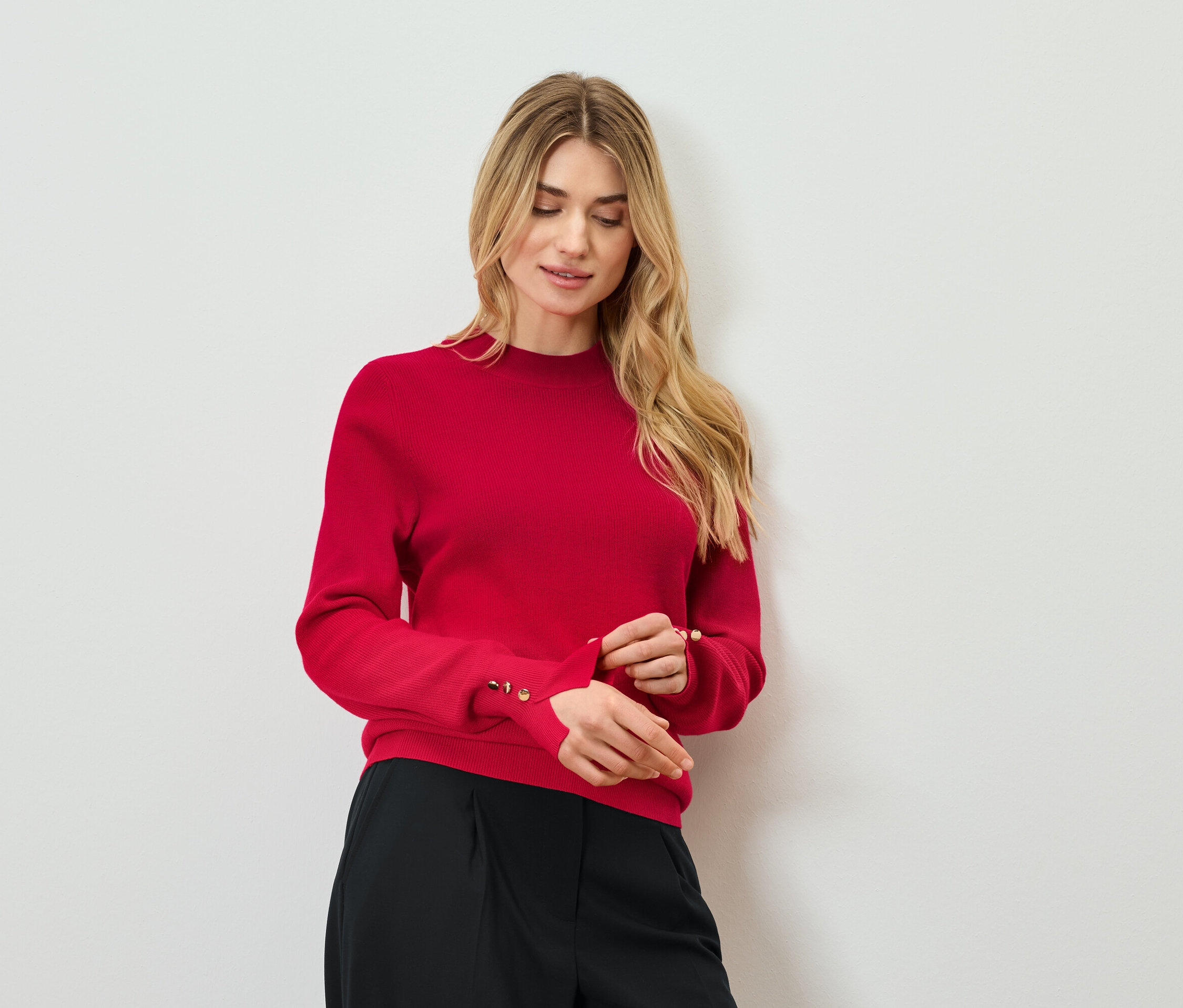 Blonde Frau justiert den Ärmel eines roten Pullovers mit Zierknöpfen. Sie trägt eine schwarze Hose.