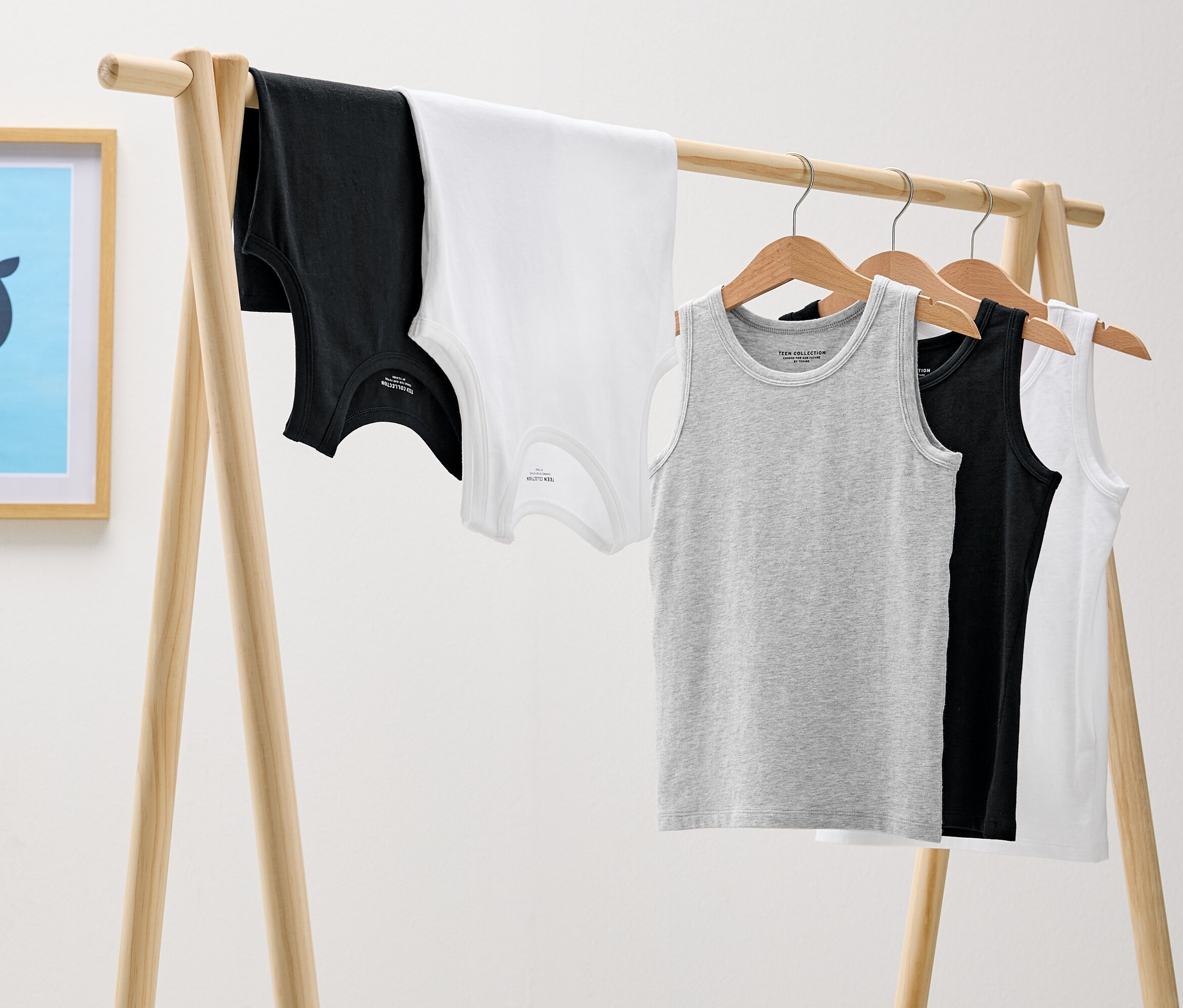 Auf einem hölzernen Kleiderständer hängen schwarze, weiße und graue Tanktops und an der Wand hängt ein Bild mit einem blauen Wal.