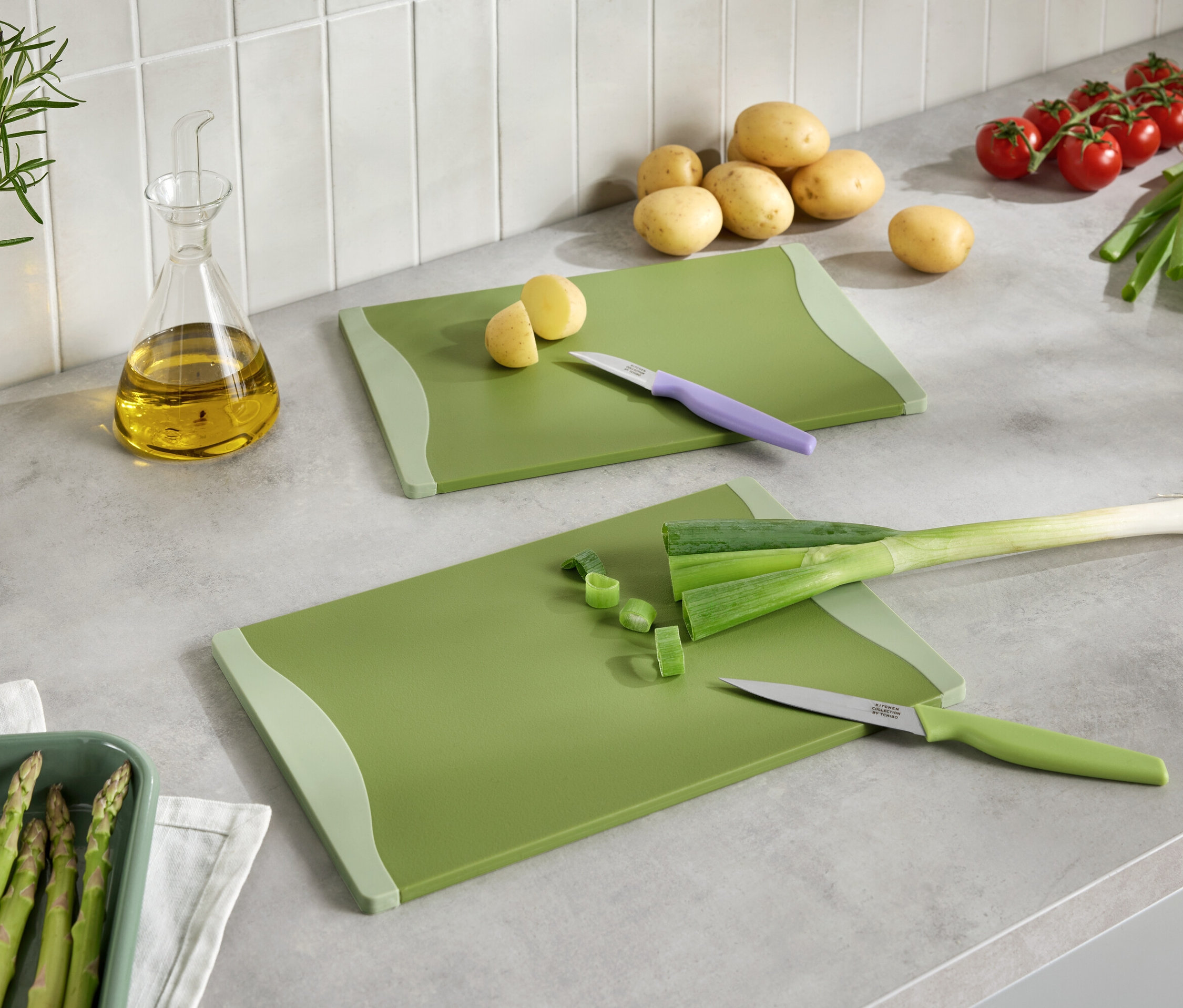 Zwei grüne Schneidebretter mit geschnittenem Gemüse, Messer, Kartoffeln, Tomaten, Lauch, Spargel und Öl auf einer Küchentheke.