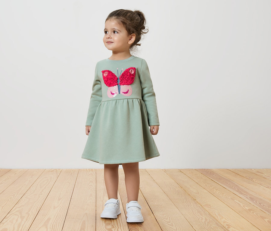 Mädchen in einem grünen Kleid mit Schmetterling steht auf einem Holzboden.