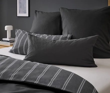 Vier graue Kissen auf einem Bett mit grau gestreifter Bettwäsche.