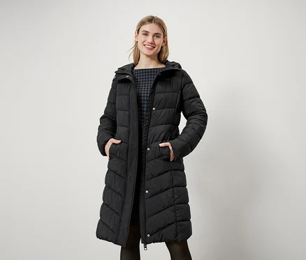 Eine Frau mit blonden Haaren steht und trägt einen langen, schwarzen Steppmantel mit Kapuze.